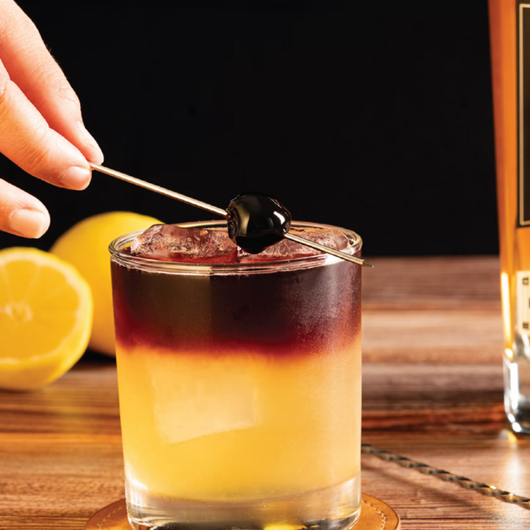 A Captain’s Sour cocktail in a rocks glass with a red wine float, lemon in the background, and garnish being placed with a bar spoon.