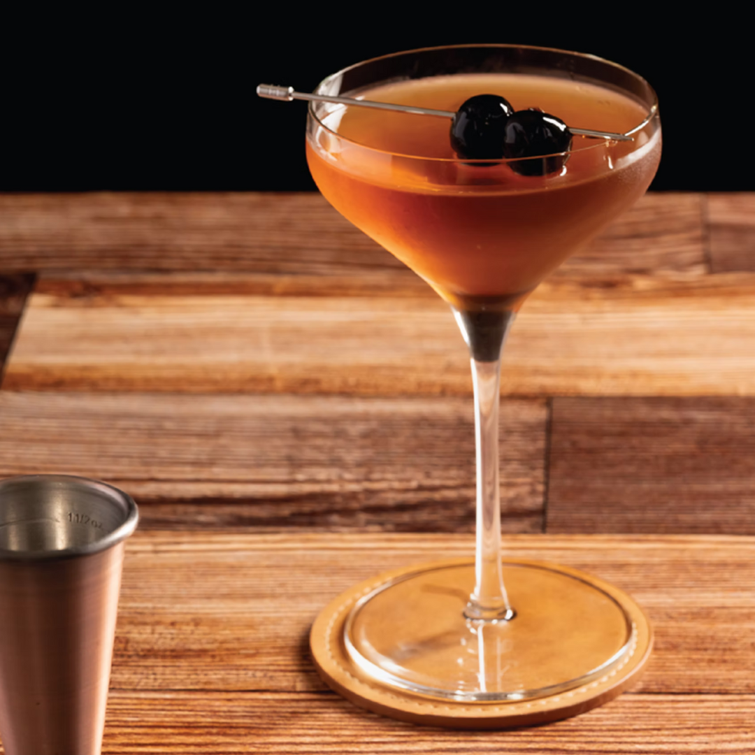 A Military Grade Manhattan cocktail served in a coupe glass with a Luxardo cherry garnish beside a Four Branches Bourbon bottle.