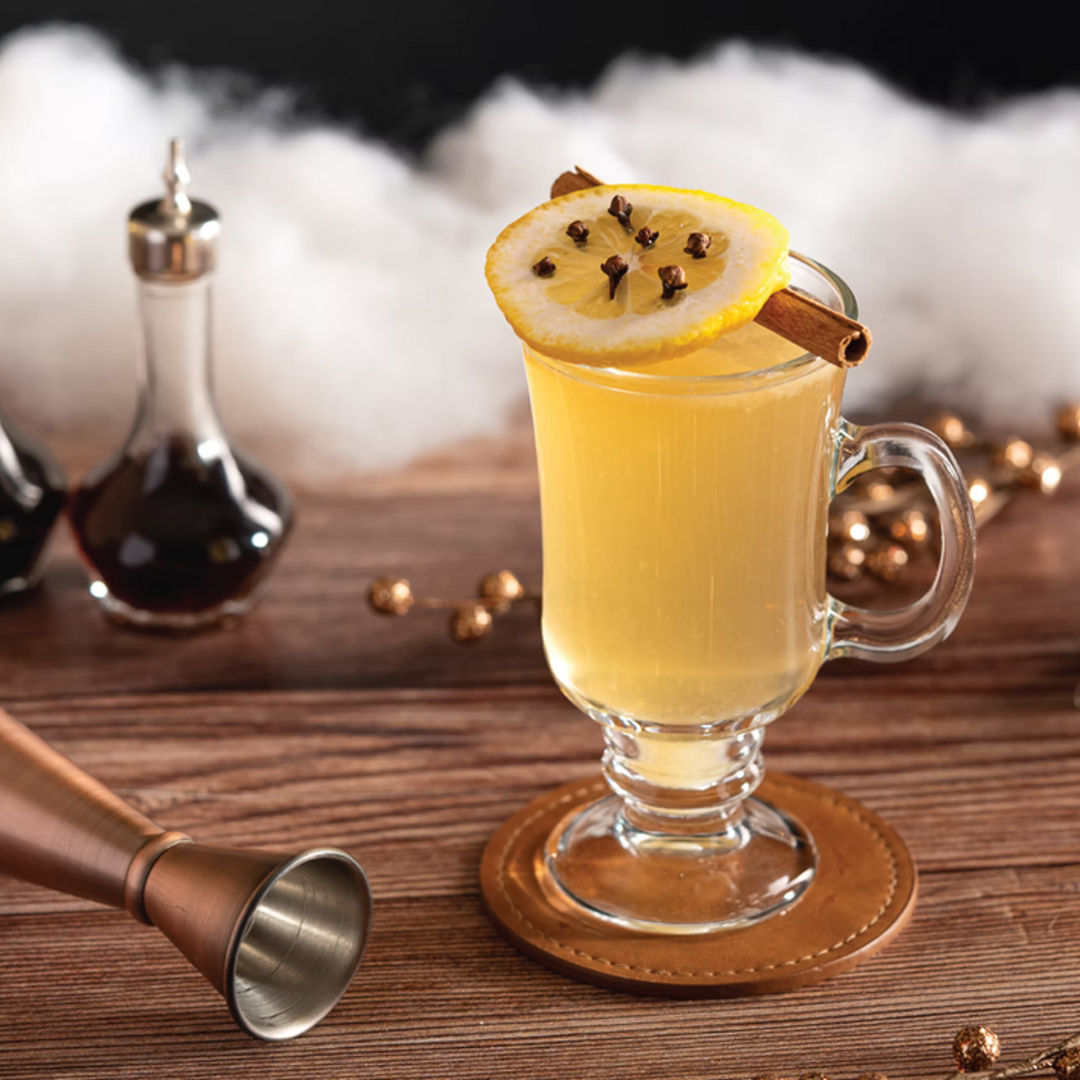 The Mount Vernon bourbon hot toddy served in a glass mug, garnished with a cloved lemon slice and cinnamon stick beside Four Branches Bourbon.