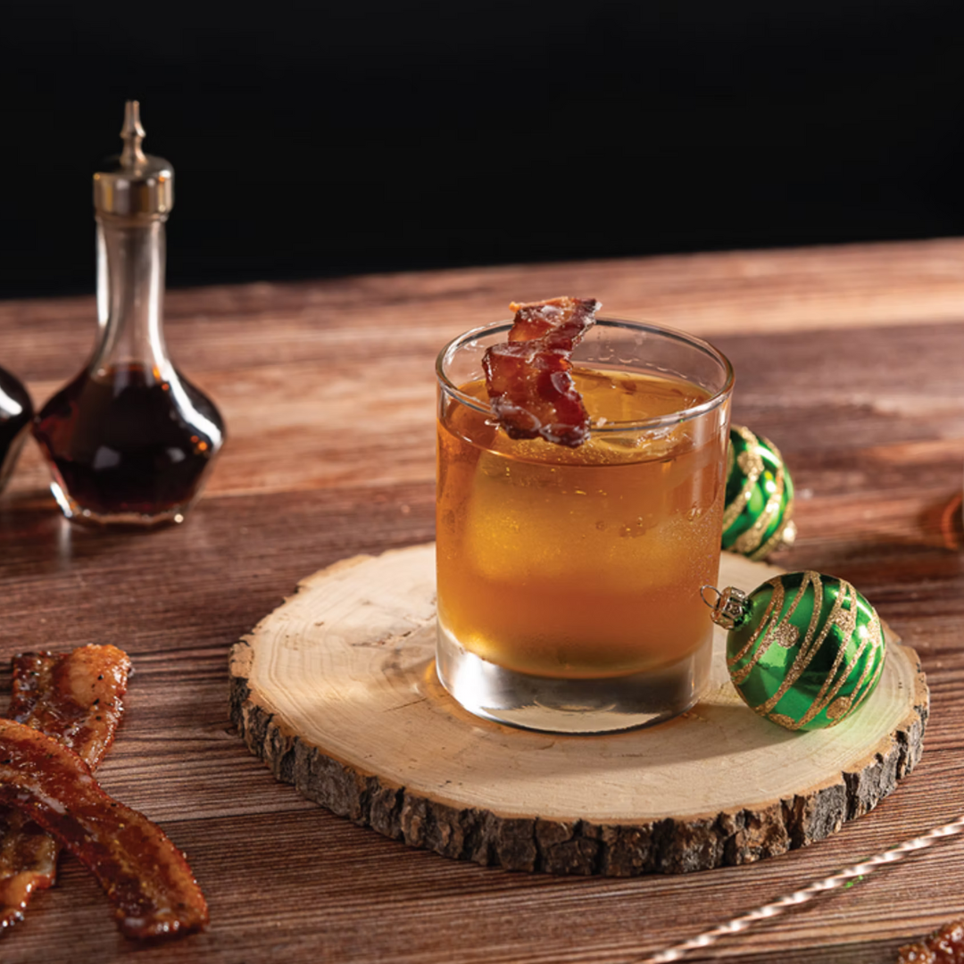 Mess Hall Old Fashioned bourbon cocktail in a rocks glass with candied bacon garnish, served on a wooden coaster with bacon strips and maple syrup nearby.